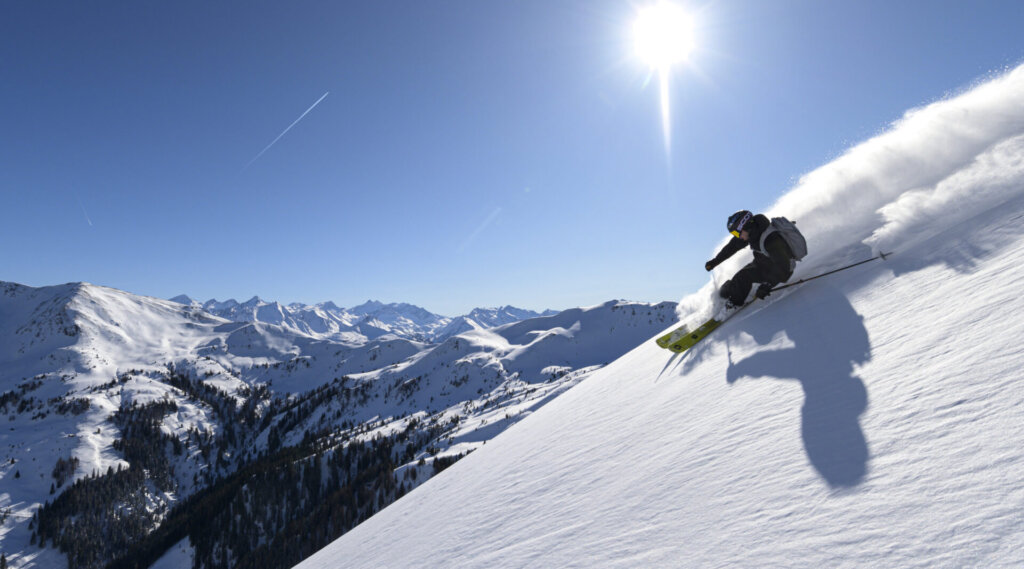 Ein Skifahrer fährt auf einem verschneiten Berghang bei strahlendem Sonnenschein und blauem Himmel bergab.