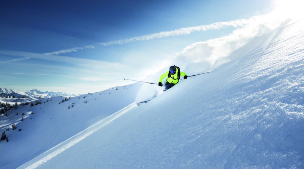 Ein Skifahrer in einer hellen Jacke fährt unter blauem Himmel einen verschneiten, sonnenbeschienenen Berghang hinunter.