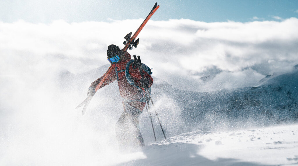 Skifahrer mit Skiern geht durch wehenden Schnee auf einem Berg, mit hellem Sonnenlicht und Wolken im Hintergrund.