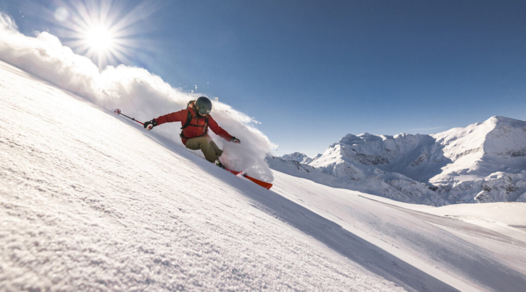 Ein Skifahrer in Rot fährt bei strahlendem Sonnenschein einen verschneiten Berghang hinunter, mit schneebedeckten Gipfeln im Hintergrund.