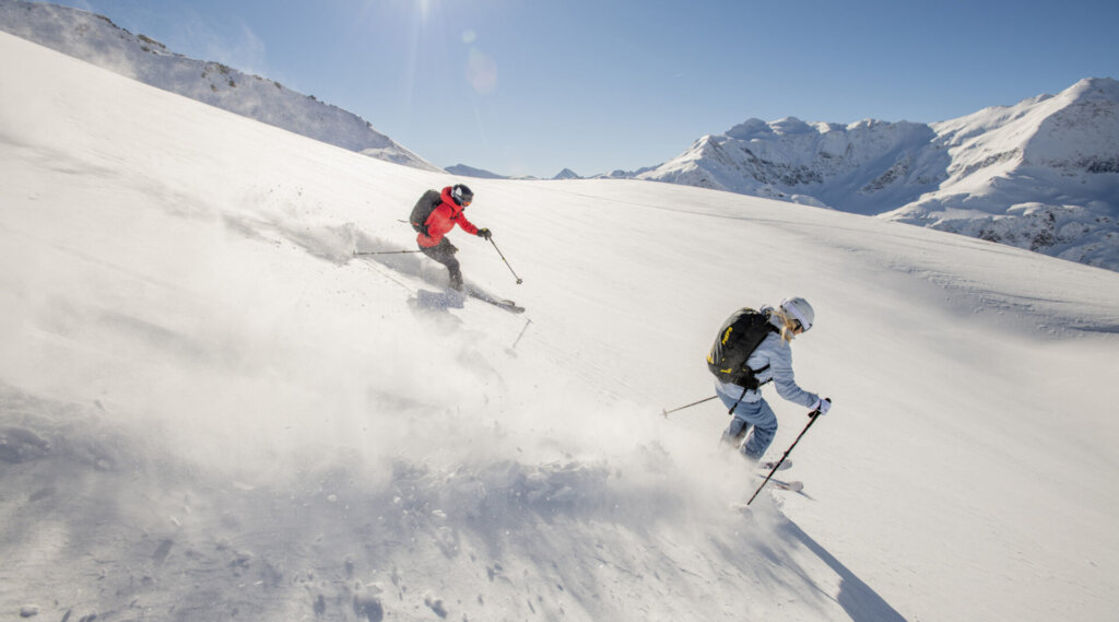 Zwei Personen fahren auf einem verschneiten Berghang unter einem hellen, klaren Himmel bergab.
