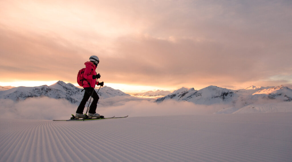 Eine Person fährt bei Sonnenaufgang auf präpariertem Schnee mit Bergen und Wolken im Hintergrund Ski.