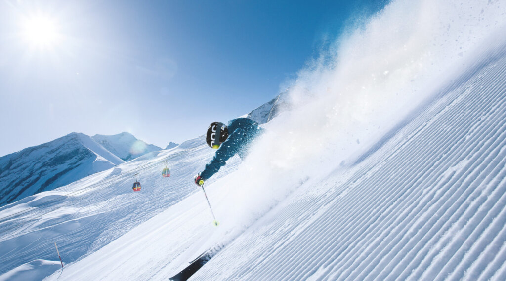 Ein Skifahrer fährt zügig einen sonnigen, verschneiten Berghang hinunter und carvt durch frischen Pulverschnee mit Bergen im Hintergrund.