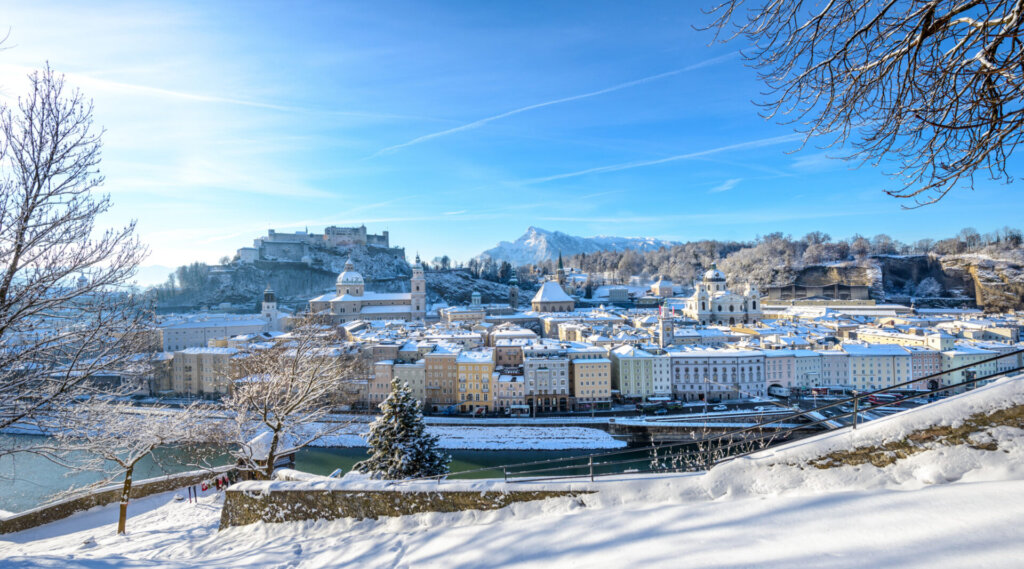 Schneebedeckte Stadtlandschaft von Salzburg, Österreich, mit Bergen und einer Festung im Hintergrund unter blauem Himmel.