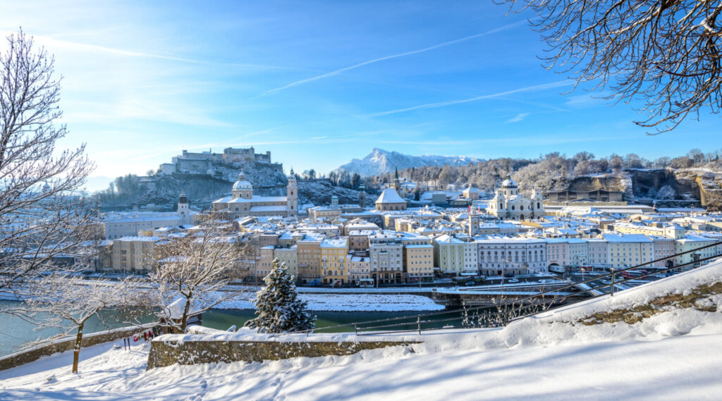 Schneebedecktes Stadtbild von Salzburg, Österreich, mit einem Fluss, historischen Gebäuden und Bergen im Winter.