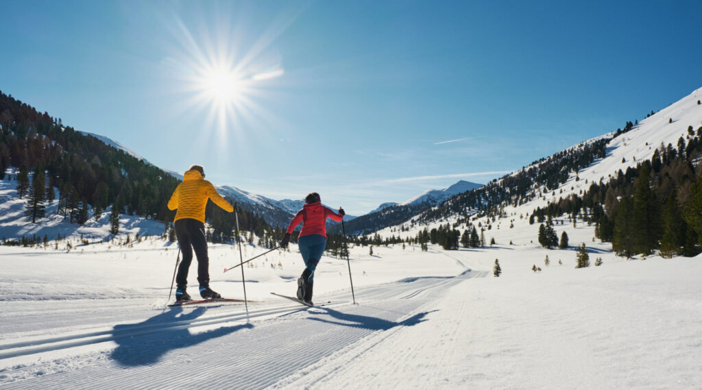 Zwei Personen beim Skilanglauf auf einer sonnigen, verschneiten Bergstrecke, umgeben von Bäumen und blauem Himmel.