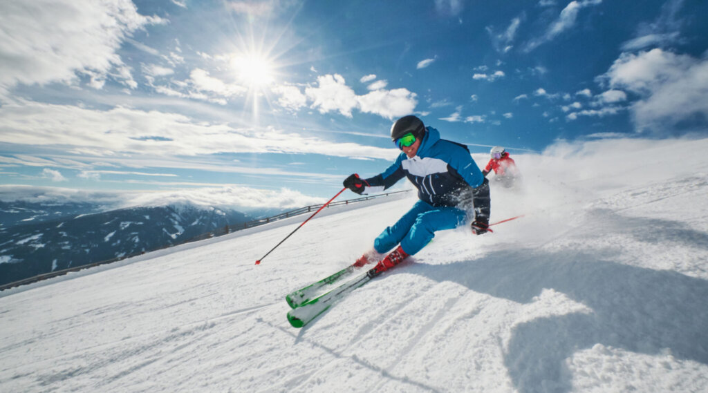 Ein Skifahrer in Blau fährt bei strahlendem Sonnenschein und teilweise bewölktem Himmel einen verschneiten Berghang hinunter.