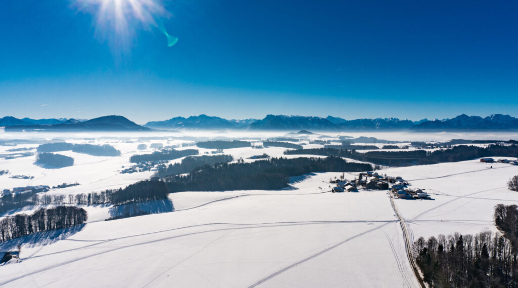 Schneebedeckte Felder mit verstreuten Bäumen und Gebäuden unter strahlender Sonne und klarem blauen Himmel, Berge in der Ferne.