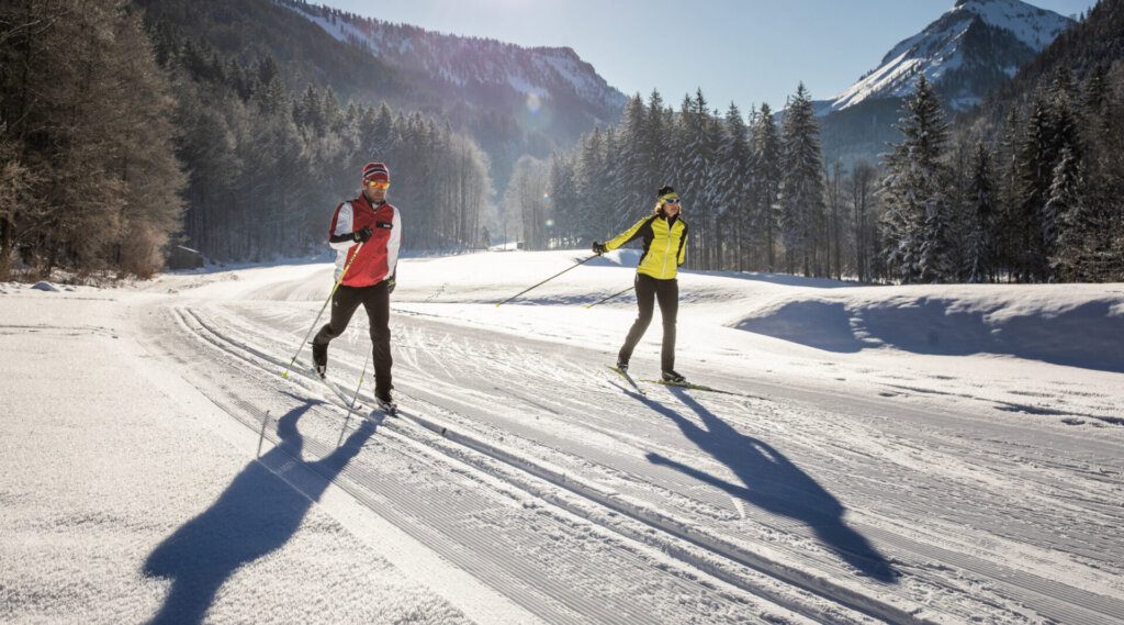 Zwei Personen beim Skilanglauf auf einer verschneiten Loipe mit Bergen und Bäumen im Hintergrund.