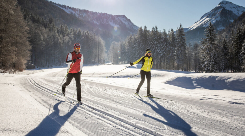 Zwei Personen beim Skilanglauf auf einer verschneiten Loipe mit Bergen und Kiefern im Hintergrund.