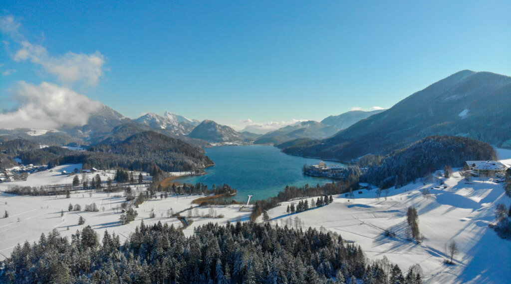 Verschneite Landschaft mit einem See und Bergen unter einem klaren blauen Himmel, umgeben von Bäumen und verstreuten Gebäuden.