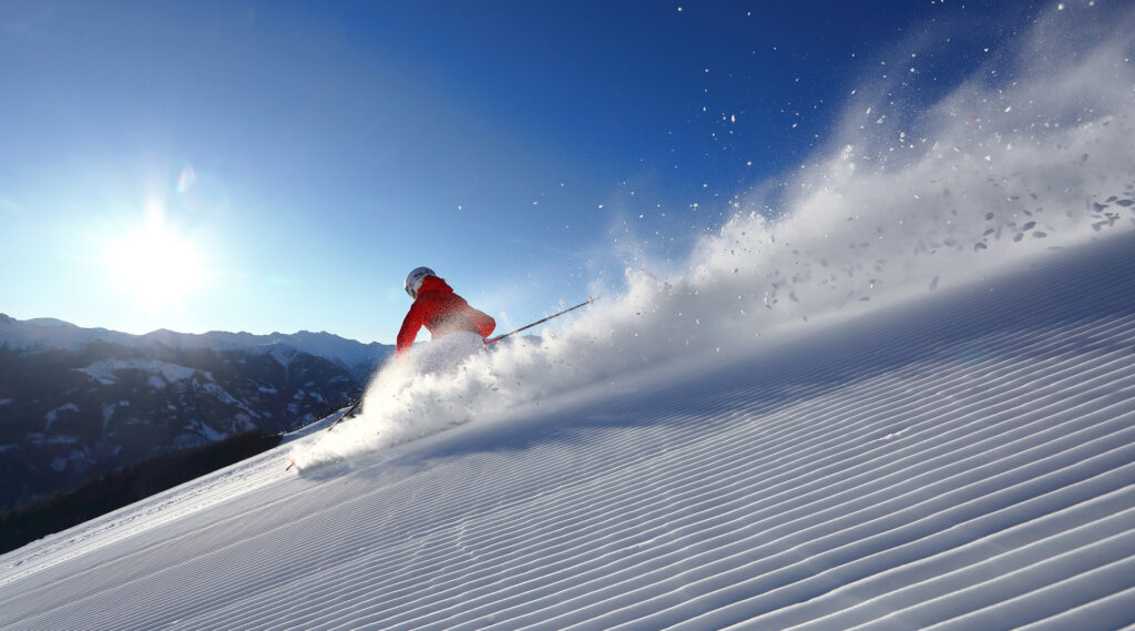 Skifahrer in roter Jacke beim Carven durch Neuschnee auf einem sonnigen Berghang mit fliegendem Pulverschnee.