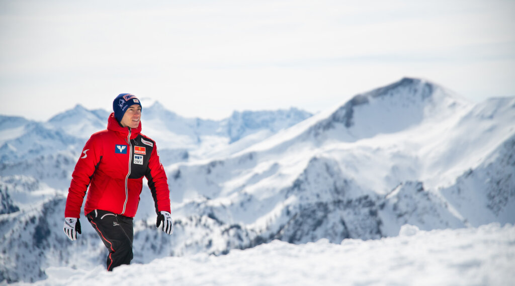 Mann in roter Jacke auf verschneitem Boden mit schneebedeckten Bergen im Hintergrund.