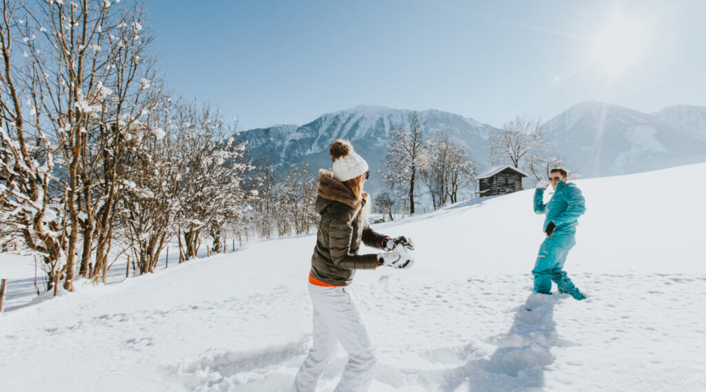 Zwei Menschen liefern sich eine Schneeballschlacht auf einem verschneiten Feld mit Bergen und heller Sonne im Hintergrund.