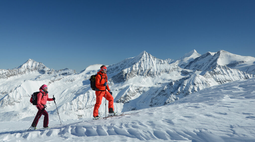 Zwei Personen in heller Winterkleidung fahren auf einem verschneiten Berg mit klarem Himmel und Alpengipfeln im Hintergrund bergauf.