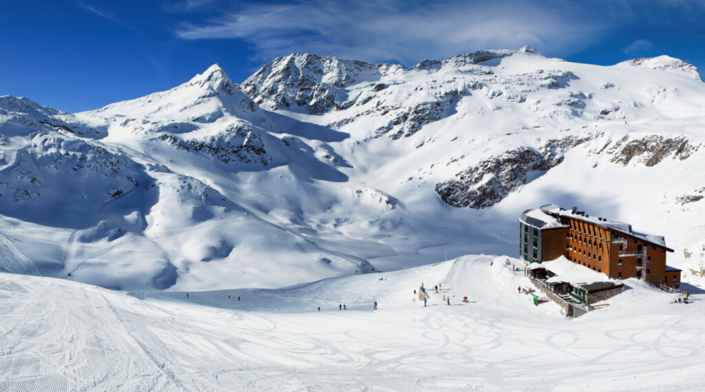 Schneebedeckte Berge mit einer Skihütte und Skifahrern auf den Pisten unter einem strahlend blauen Himmel.