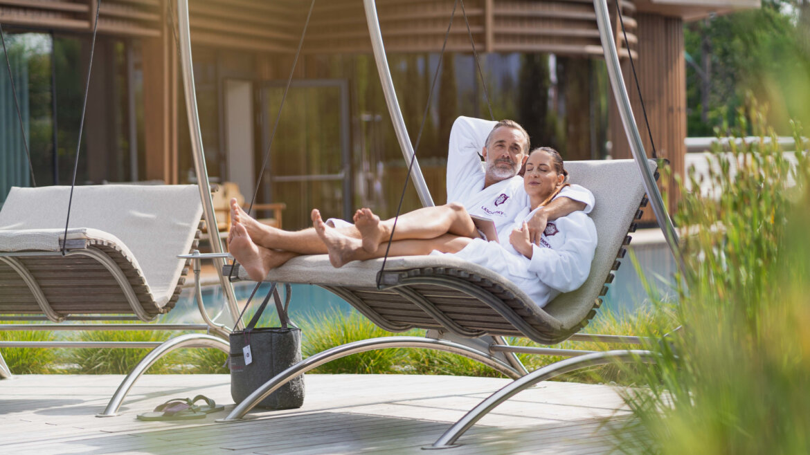 A couple in white dressing gowns relax together on an outdoor lounger swing by a pool at a modern spa. (Enlarged view)