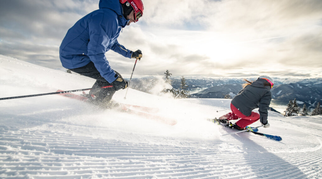 Zwei Skifahrer mit Helmen und Winterausrüstung fahren auf einem verschneiten Berg bei bewölktem Himmel bergab.