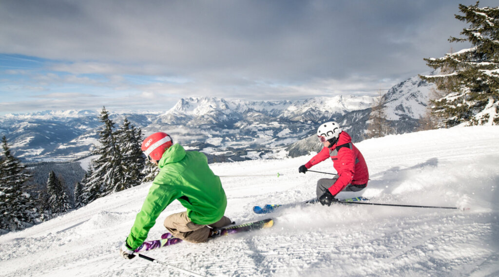 Zwei Personen fahren auf Skiern einen verschneiten Berghang hinunter, mit Bäumen und Bergen im Hintergrund.