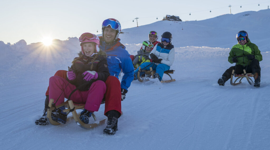 Fünf Menschen rodeln einen verschneiten Hügel hinunter, lächeln und genießen die Wintersonne.