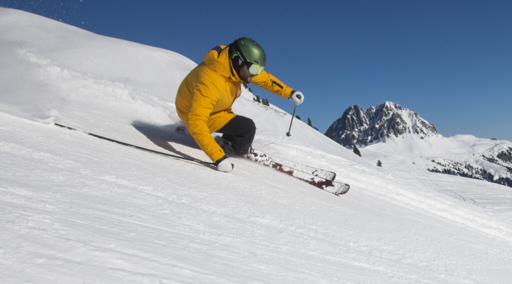 Person in gelber Jacke beim Skifahren auf einer verschneiten Piste mit Bergen im Hintergrund unter einem klaren Himmel.