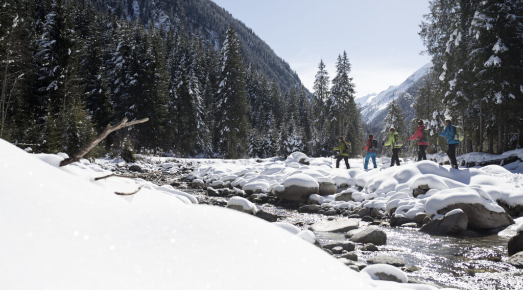 Eine Gruppe von Menschen geht an einem verschneiten Flussufer entlang, umgeben von schneebedeckten Bäumen und Bergen.