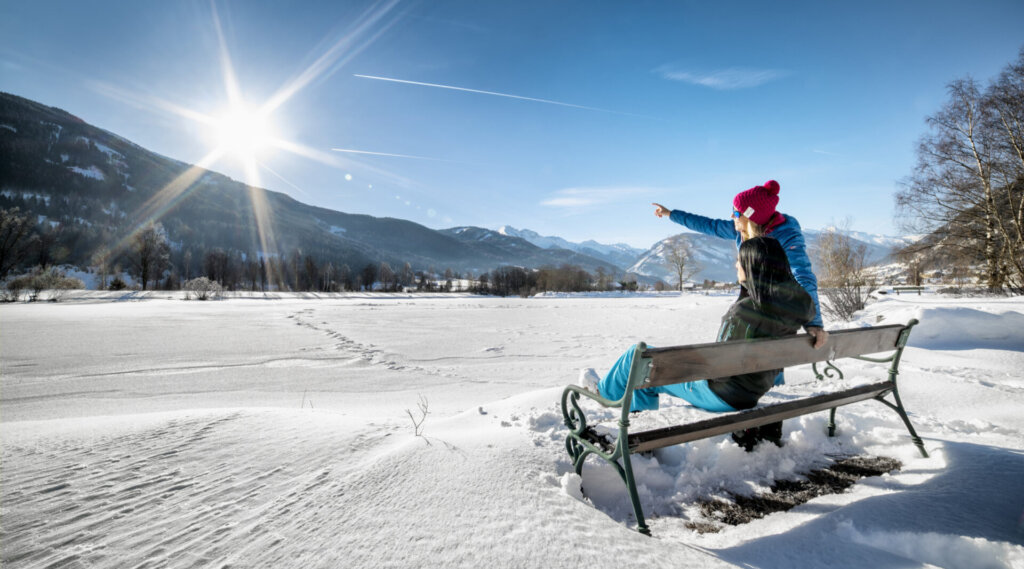 Zwei Menschen sitzen auf einer verschneiten Bank; einer zeigt auf die sonnenbeschienenen Berge unter einem strahlend blauen Himmel.