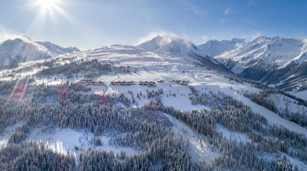 Luftaufnahme einer verschneiten Berglandschaft mit Sonnenschein über Kiefernwäldern und verstreuten Hütten.