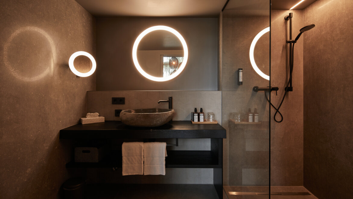 Modern bathroom with round backlit mirrors, stone basin, black worktop, and glass shower enclosure. (Enlarged view)