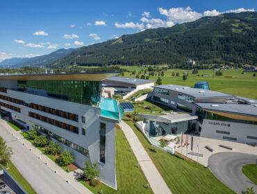 Modern spa resort buildings with green lawns, mountains, and blue sky in the background.