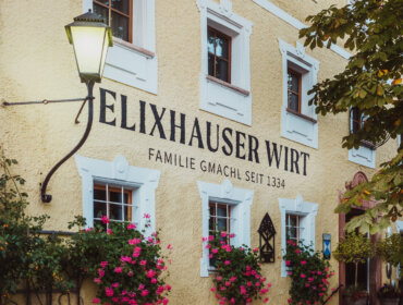 Yellow building with flower boxes and sign reading “Elixhauser Wirt Familie Gmachl seit 1334” on the wall.