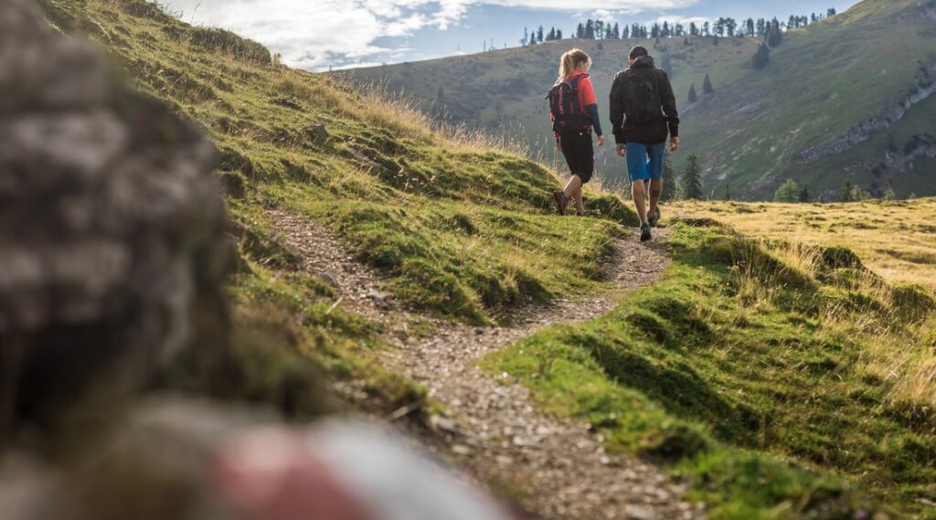 Hiking at Postalm (c) Michael Groessinger Two people with rucksacks hiking on a winding path through grassy hills under a partly cloudy sky. (Enlarged view)