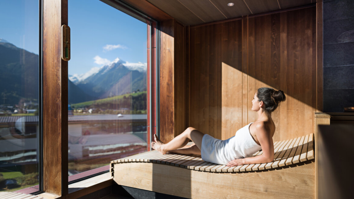 © TAUERN SPA Zell am See-Kaprun A woman relaxes in a sauna, wrapped in a towel, looking out at snowy mountains through a large window. (Enlarged view)