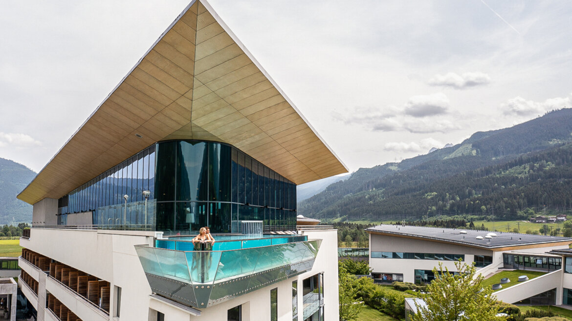 © TAUERN SPA Zell am See-Kaprun A person stands on a glass balcony of a modern building with mountains and greenery in the background. (Enlarged view)