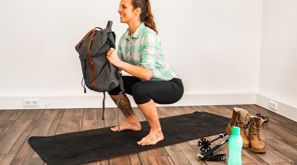 Woman in a checked shirt squatting on a mat, holding a rucksack, with boots, pole, and bottle nearby.