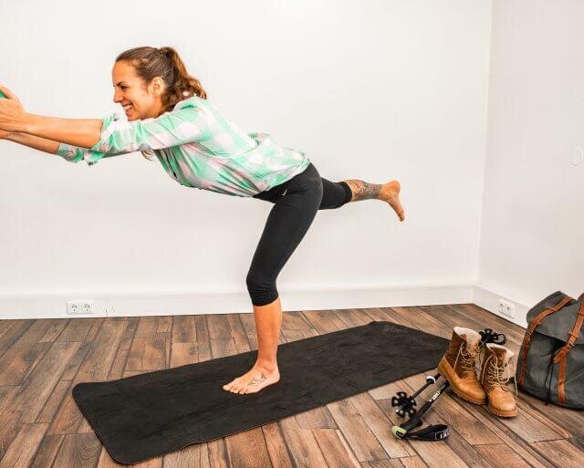 Woman balancing on one leg on a yoga mat indoors, with hiking kit and boots nearby on the floor.