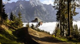 Two mountain bikers jump on a forest trail with mountains and clouds in the background.