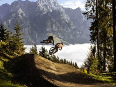 Two mountain bikers jump on a forest trail with mountains and clouds in the background.