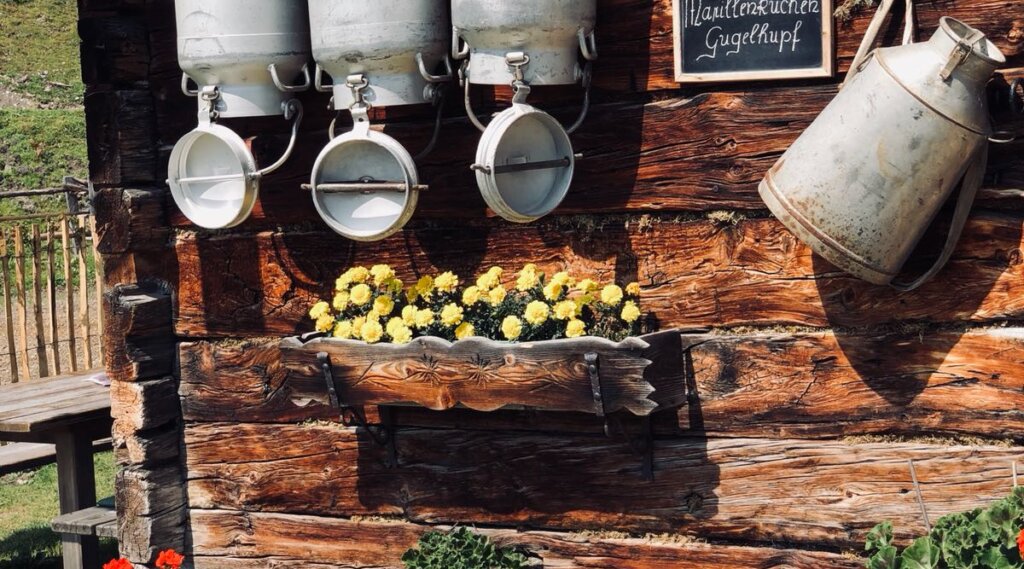 Milk churns at Tauernkarleitenalm (c) Untersulzbergalm Metal milk churns and yellow flowers on a rustic wooden wall, with a chalkboard sign above. (Enlarged view)