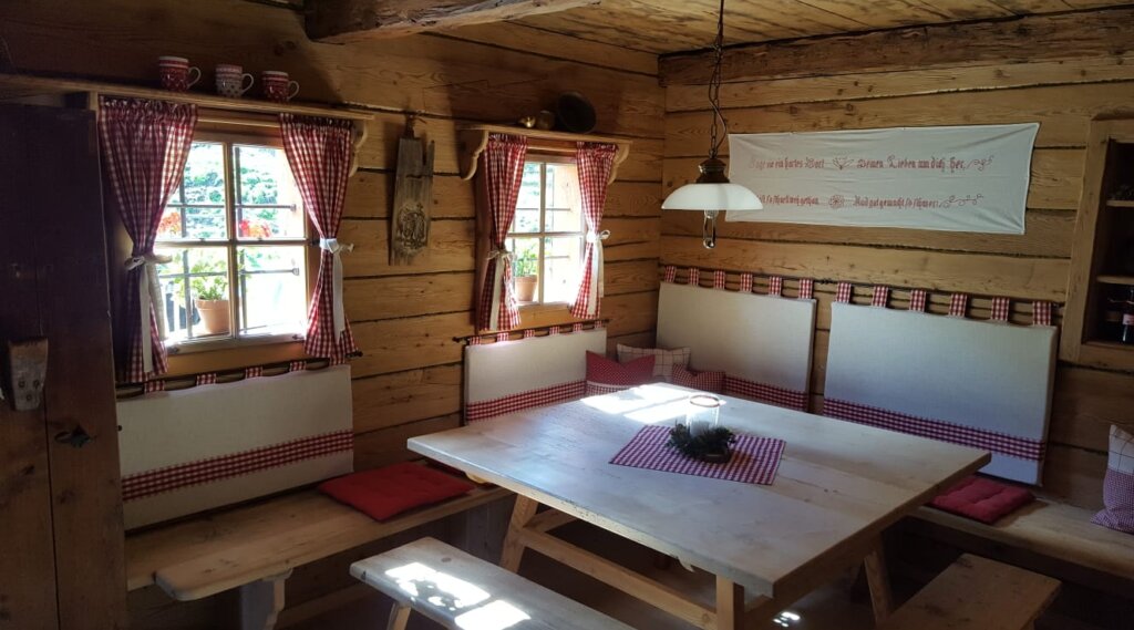 Tauernkarleitenalm indoor (c) Untersulzberghof Cosy rustic dining nook with wooden walls, red chequered curtains, and a white table with benches. (Enlarged view)