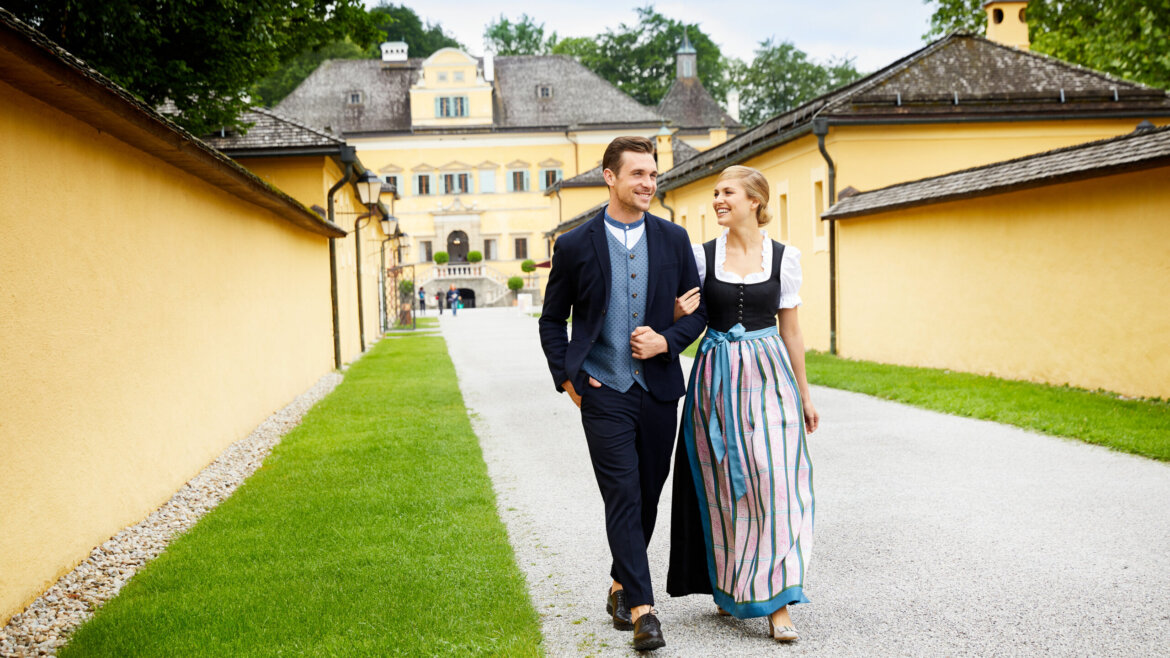 A smiling couple in traditional Austrian clothing walk arm in arm on a path by a yellow historic building.