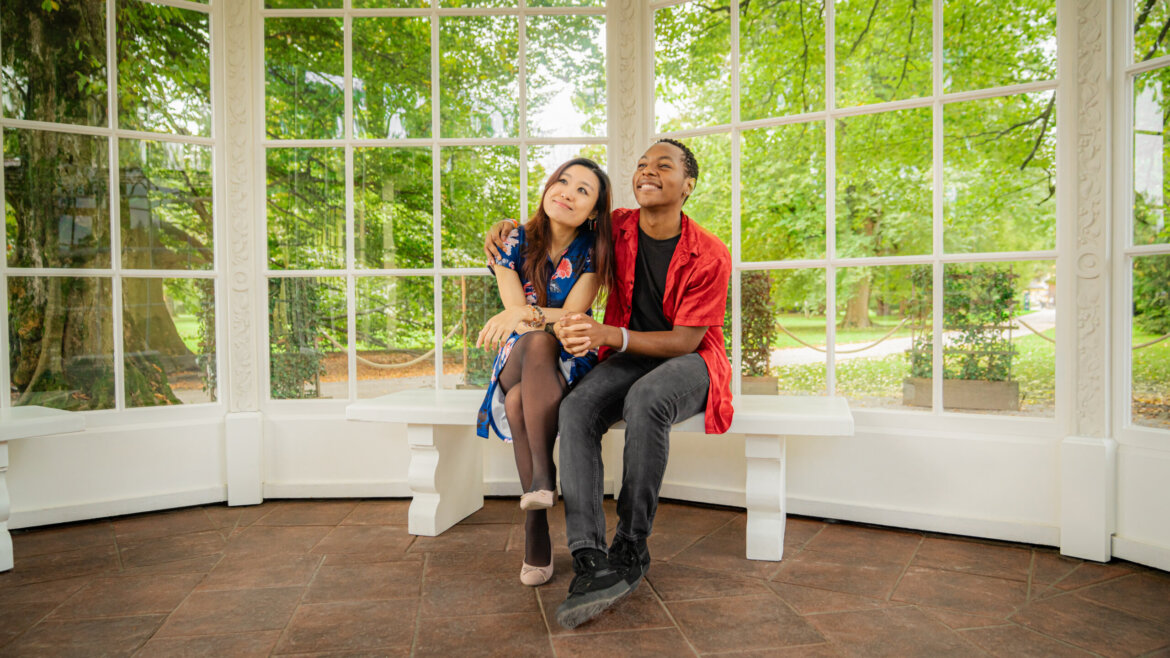 Two people sit close together on a bench in a bright gazebo, smiling, with green trees visible through large windows.