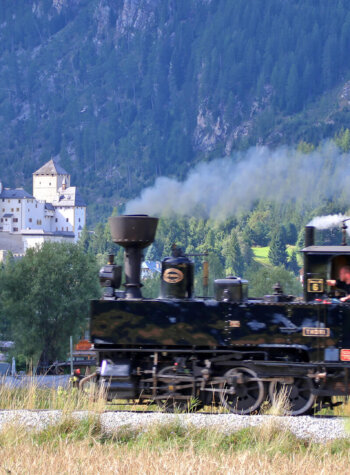 A vintage steam train passes through a green valley with a white castle and forested hills in the background.