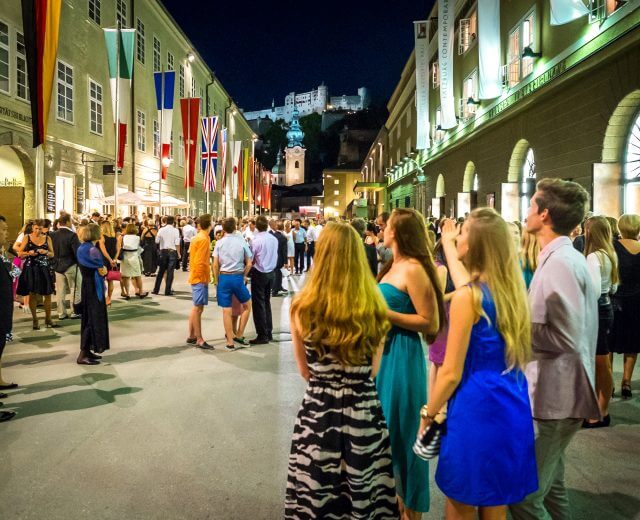 Nyári ruhás tömeg gyűlik össze éjszaka, zászlókat lengetve egy vár közelében a Salzburgi Ünnepi Játékok alatt.