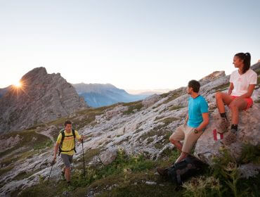 Wanderung zur Passauer Hütte Három túrázó pihen egy sziklás hegyoldalon napfelkeltekor, élvezik a kilátást és megosztják biztonsági tanácsok.