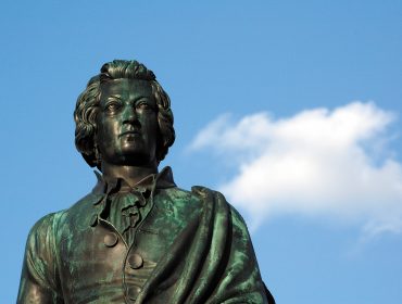 Mozart göndör hajú bronzszobra áll a kék égbolton, amelyen egyetlen fehér felhő van.