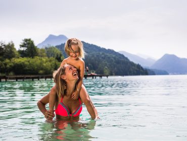 Rózsaszín bikinis nő mosolygó gyermeket visz a tó- és strandfürdők tójában, mögötte hegyek.