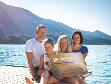 Egy négytagú család ül mosolyogva egy tóparti stégen, a háttérben hegyekkel, kezében egy táblával.