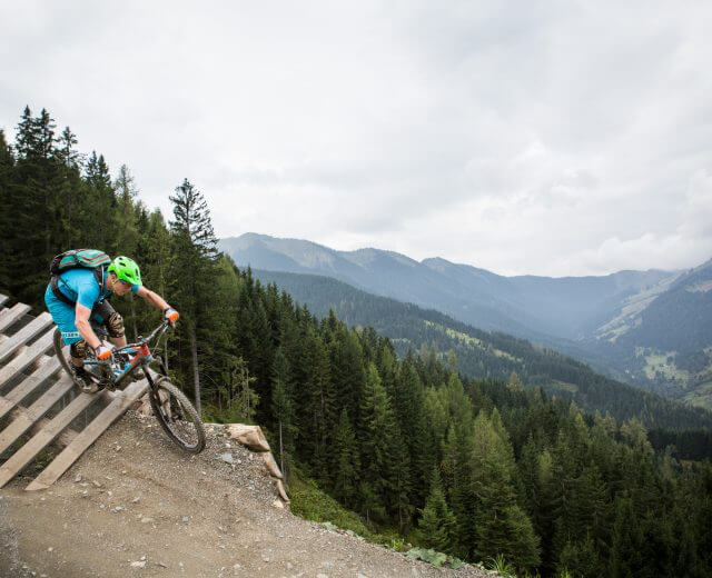 Kék ruhás hegyi kerékpáros hajt le egy fából készült rámpán a bringaparkokban, mögötte erdős dombok és felhős hegycsúcsok.