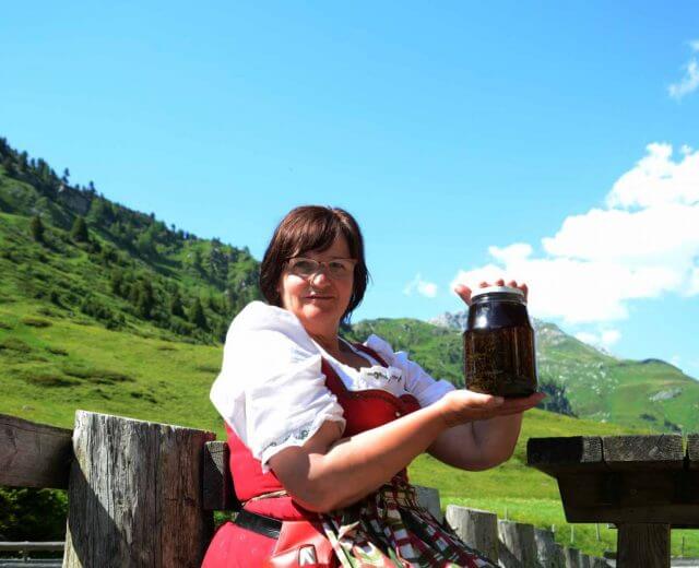 Egy hagyományos ruhás nő tart egy korsót, a gyógymódok ihlette kint a szabadban, mögötte zöld dombok és kék égbolt.